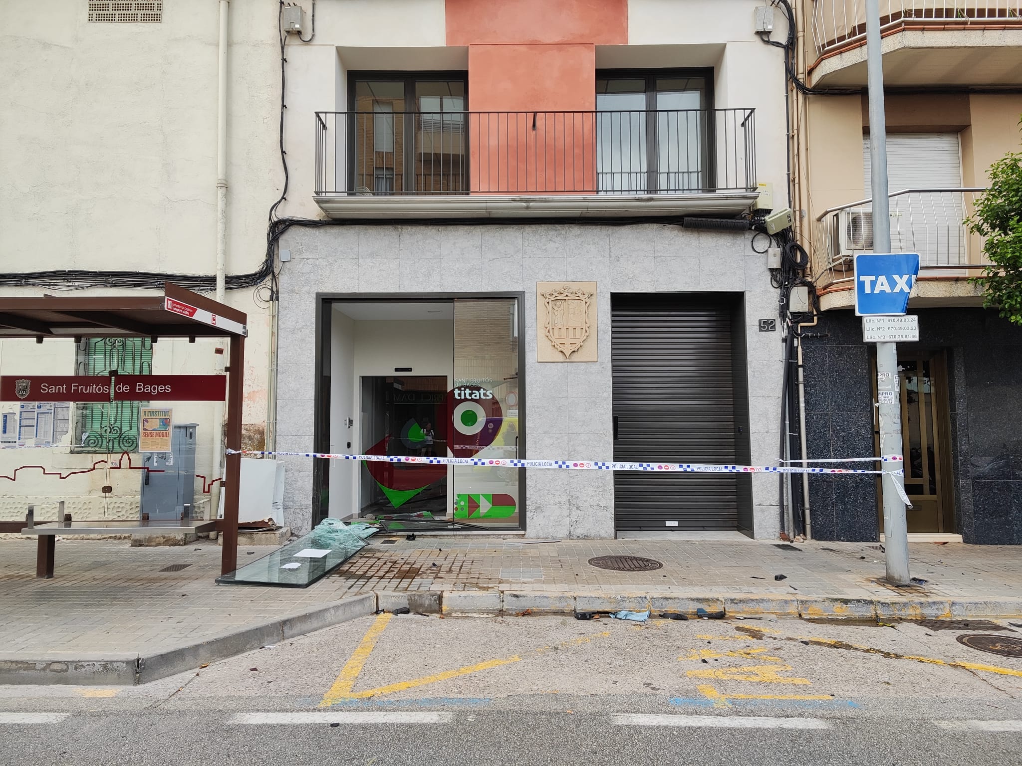 Façana del Casal d’Entitats de Sant Fruitós de Bages amb la porta principal trencada després que un cotxe hagi envaït la vorera i impactat contra l’edifici. La zona està acordonada amb cinta policial i s’observa la marquesina de la parada d’autobús i un senyal de taxi.